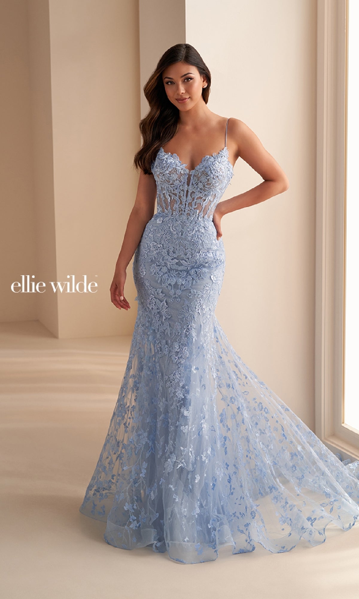A woman wearing the Ellie Wilde EW36008, a long embroidered prom dress in light blue with thin straps and lace details, stands indoors near a window. The image displays